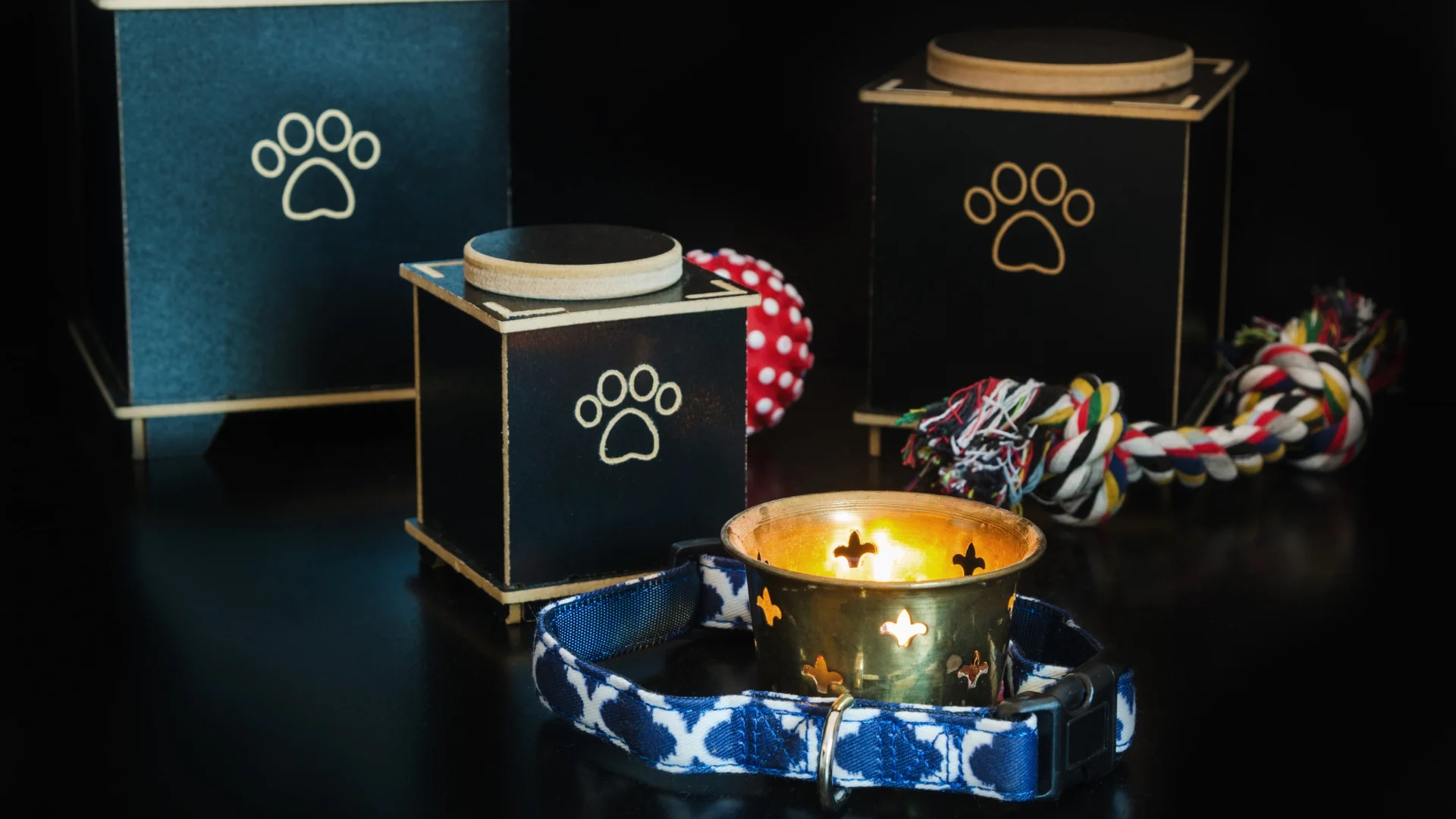 A lit candle, dog collar, and toy placed in front of pet cremation urns with paw prints on them.